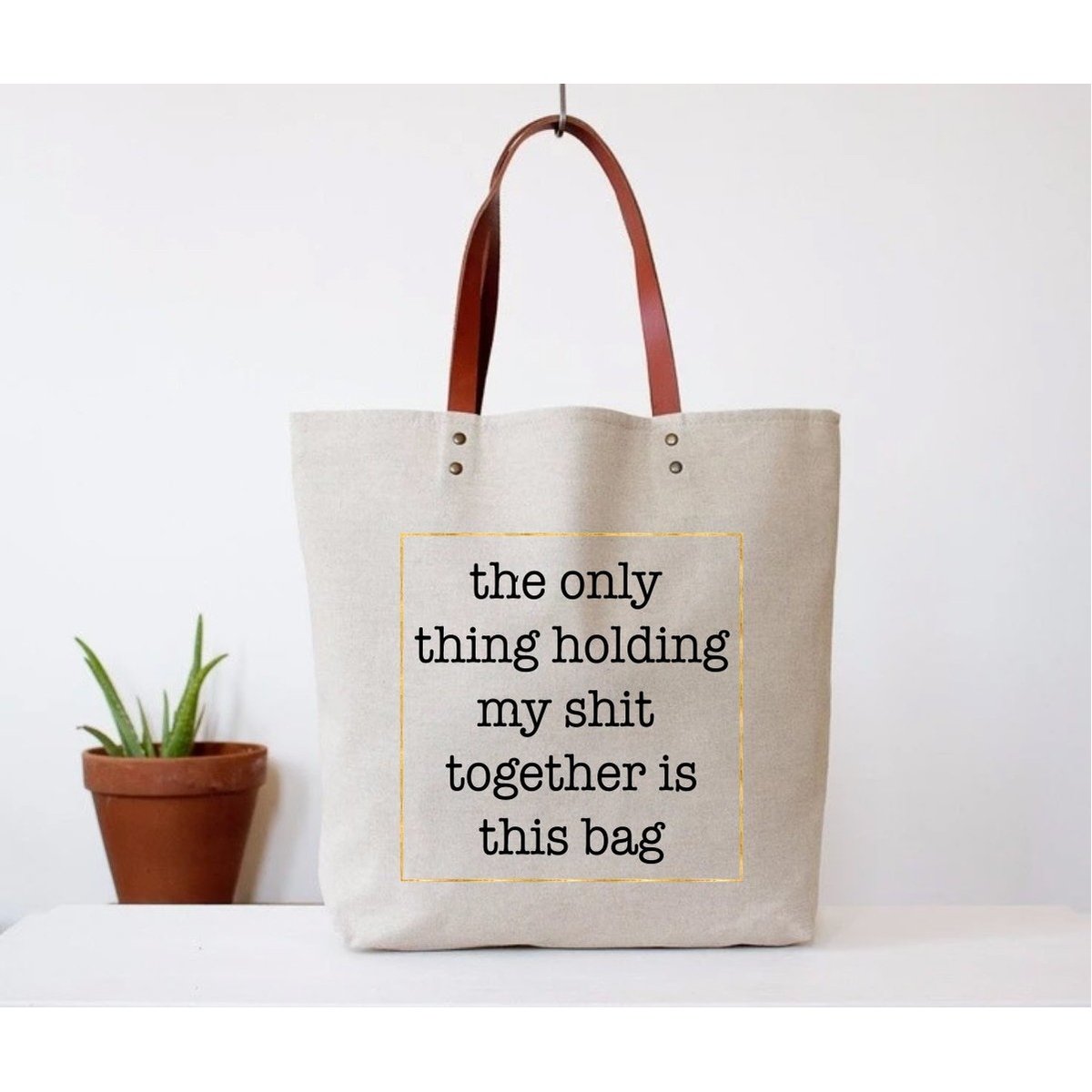 Canvas tote bag with faux leather straps that says "the only thing holding my shit together is this bag"