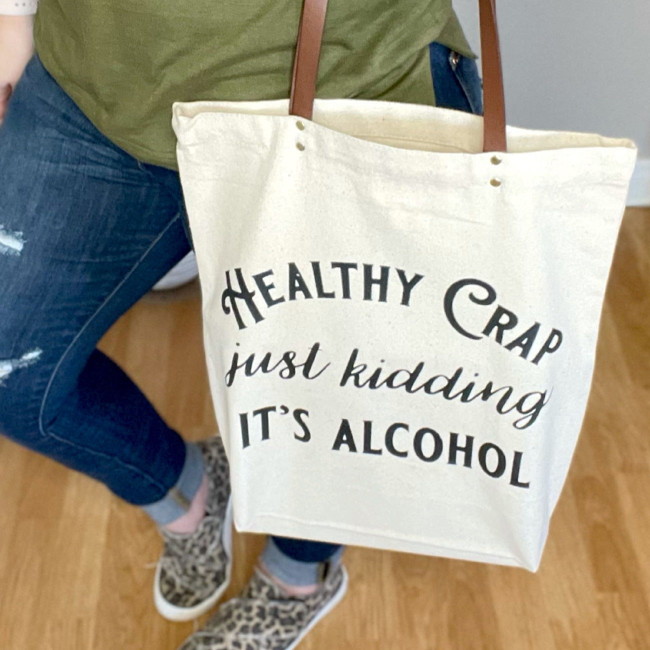 Woman holding a canvas tote bag that says "Healthy Crap - Just kidding it's alcohol"